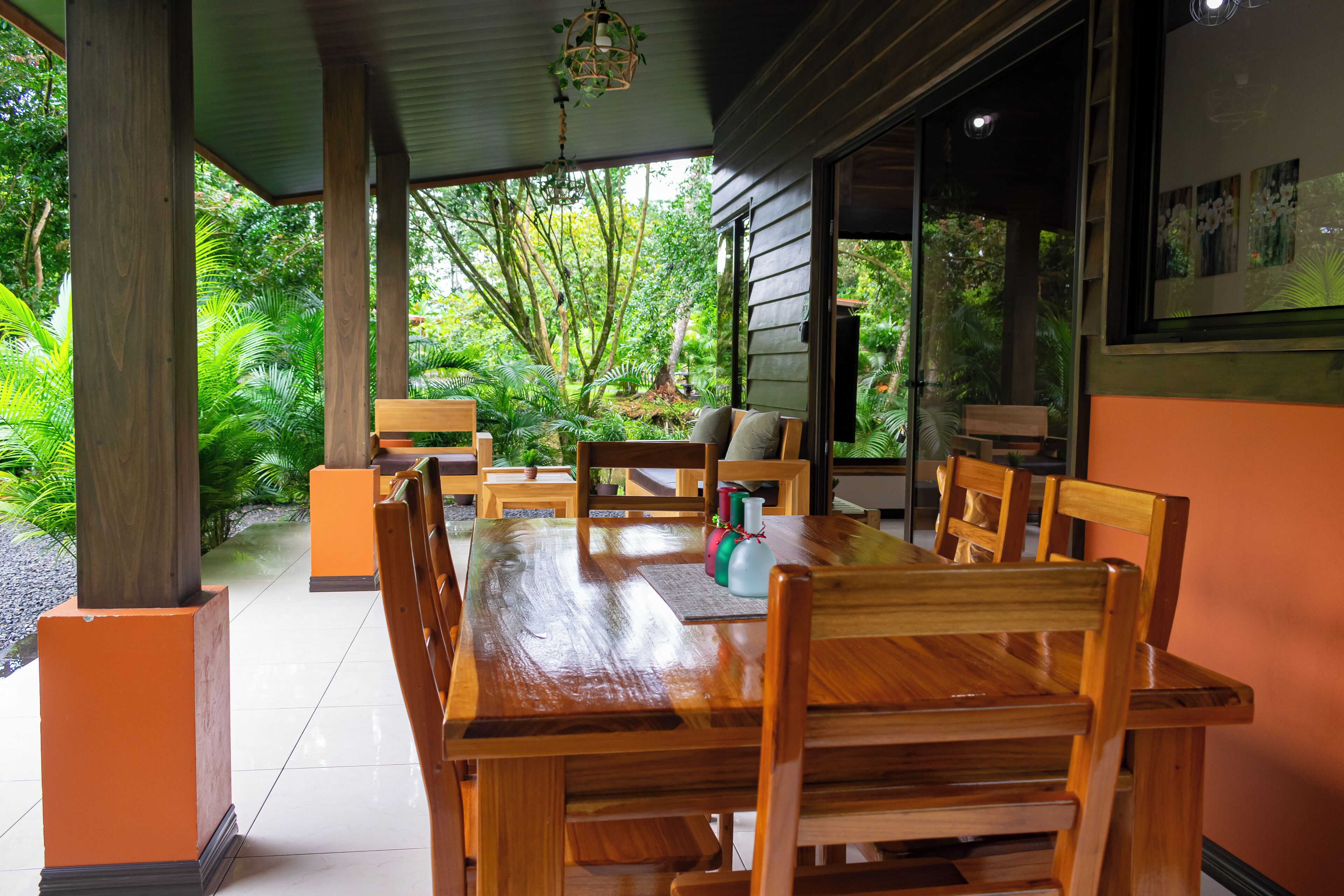 An outdoor wooden dining area with a table and chairs, surrounded by greenery and a glass door leading to a lush garden.