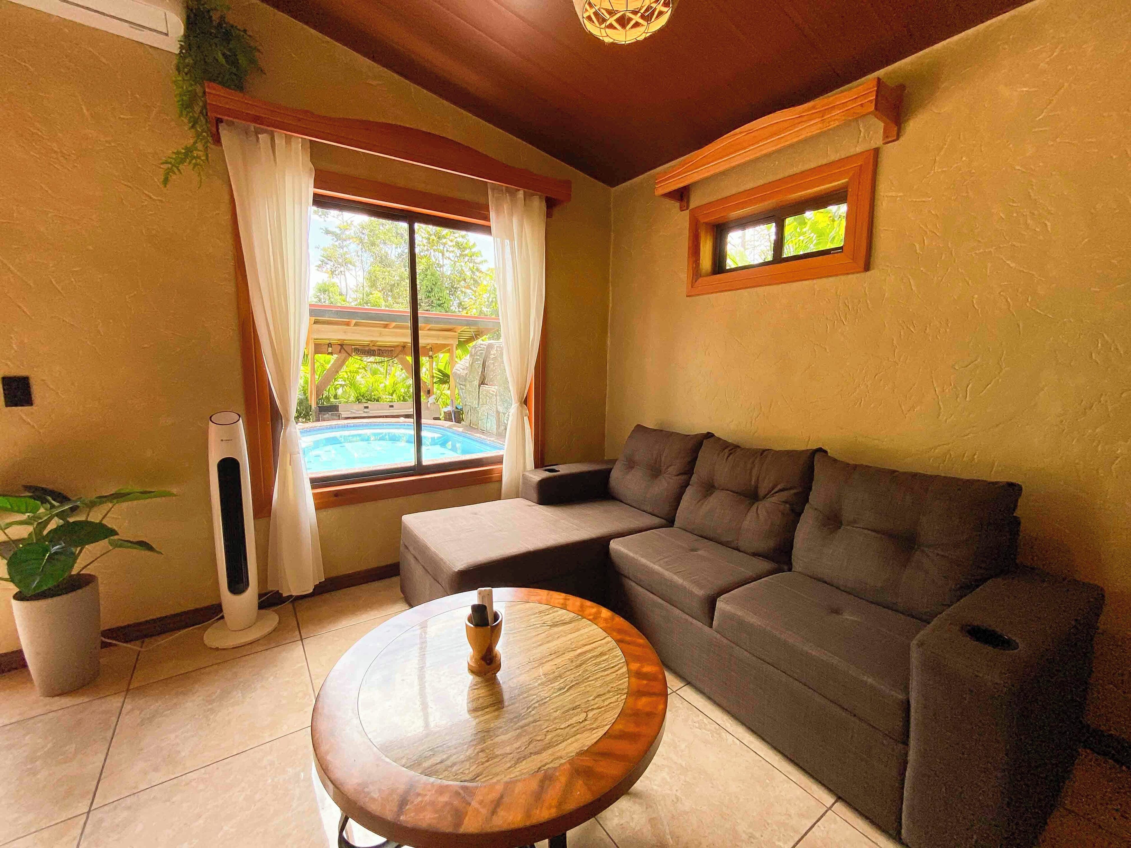 A living room with a gray sofa, a wooden coffee table, a massage chair, a large window with curtains, a small window, and a ceiling fan.