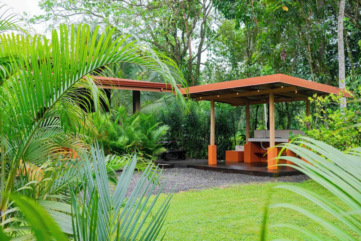 Un espacio de descanso al aire libre con techo, estructura de madera y un banco, rodeado de abundante vegetación y un césped cuidado.