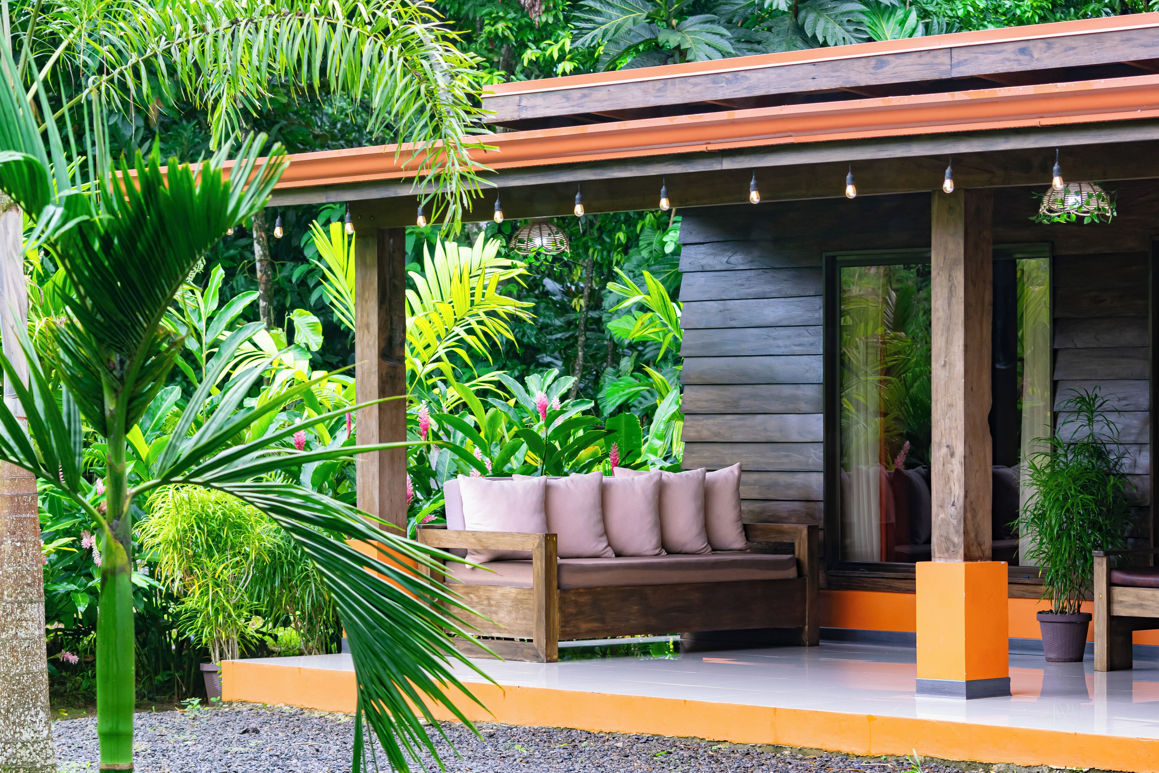 Eine Holzterrasse mit gepolsterten Sitzgelegenheiten, umgeben von üppigen tropischen Pflanzen und einer Holz-Pergola mit Lichterketten.