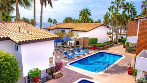 Un área de piscina con sillones de descanso y una piscina pequeña, rodeada de palmeras y edificios con tejados de tejas.