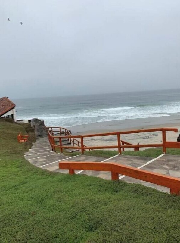 A coastal walkway with steps leading to a beach, a wooden railing, and a building with a red roof in the background. - Cozy condo 2 minutes from the beach at a tennis resort gated community (Playas de Rosarito)
