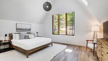 A bedroom with a bed, a nightstand, a lamp, a dresser, and a large window with a view of trees.