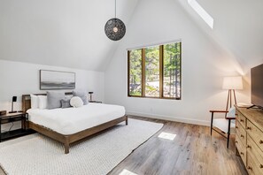 Un dormitorio con cama, mesita de noche, lámpara, cómoda y un ventanal con vistas a árboles.