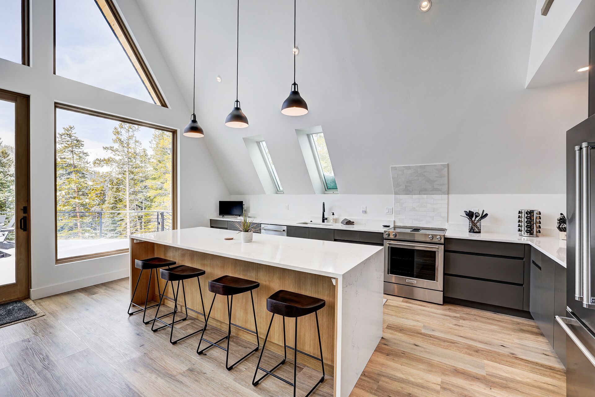 A modern kitchen with a large island, bar stools, a built-in oven, and a view of trees outside.