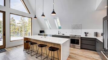 A modern kitchen with a large island, bar stools, a built-in oven, and a view of trees outside.