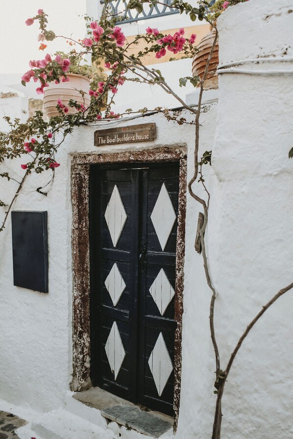 A white building with a black door featuring diamond-shaped white patterns, a sign above the door, and pink flowers growing on the wall.