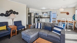 Ein gemütlicher Wohnraum mit einem grauen Ecksofa, einem blauen Sessel und einem Holztisch. Im Hintergrund ist eine Küche mit weißen Schränken und Edelstahlgeräten zu sehen.