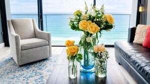 A modern living room with a large window overlooking the ocean, a grey armchair, a blue sofa, and a table with floral arrangements. - Ocean View Luxury Condo (San Diego)