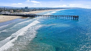 A pier extending into the ocean with clear blue water, a sandy beach, and a distant cityscape. - Ocean View Luxury Condo (San Diego)
