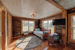 A bedroom with a wooden ceiling, a bed with a headboard, a television mounted on the wall, and a large window.