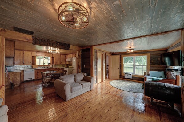 A spacious cabin interior with a wooden floor, a dining area, a living room with a sofa, and a kitchen with a stove and refrigerator.