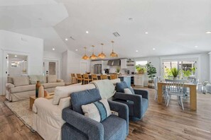 Uma sala de estar espaçosa com um conjunto de sofás, uma área de jantar com mesa e cadeiras, e uma cozinha ao fundo.