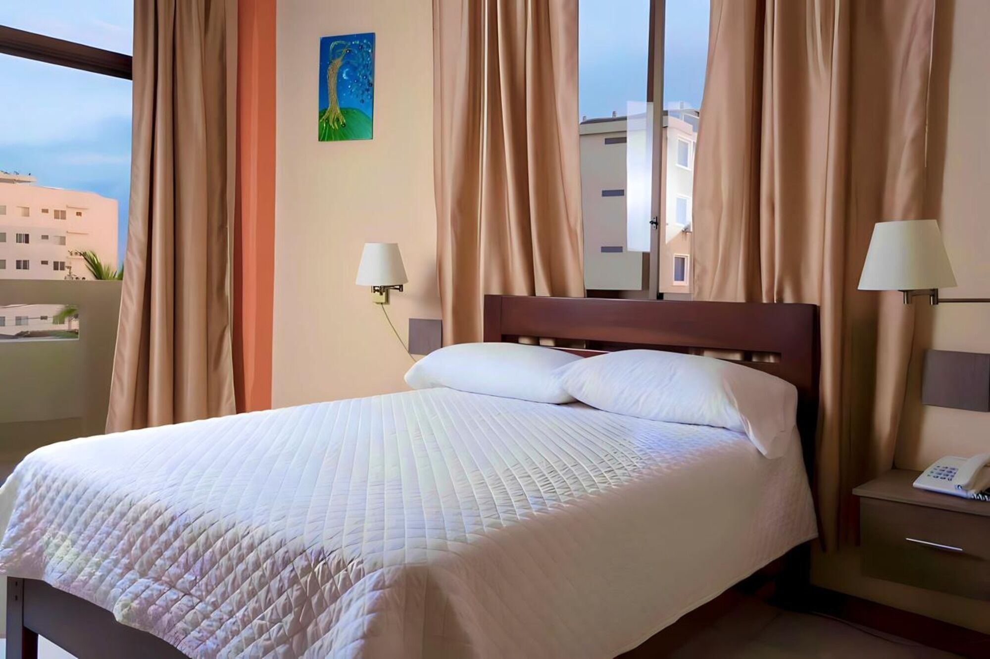 A hotel room with a bed, two bedside lamps, a phone on the nightstand, and a painting on the wall.