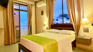 Habitación de hotel con cama, mesita de noche, lámpara, equipo de aire acondicionado y vista a edificios por la ventana.