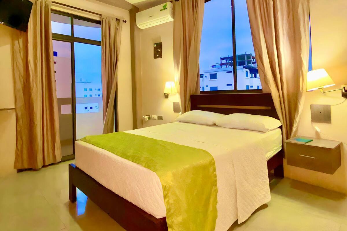 Habitació d'hotel amb un llit, una taula de nit, una làmpada, un aparell d'aire condicionat i vista a edificis a través de la finestra.
