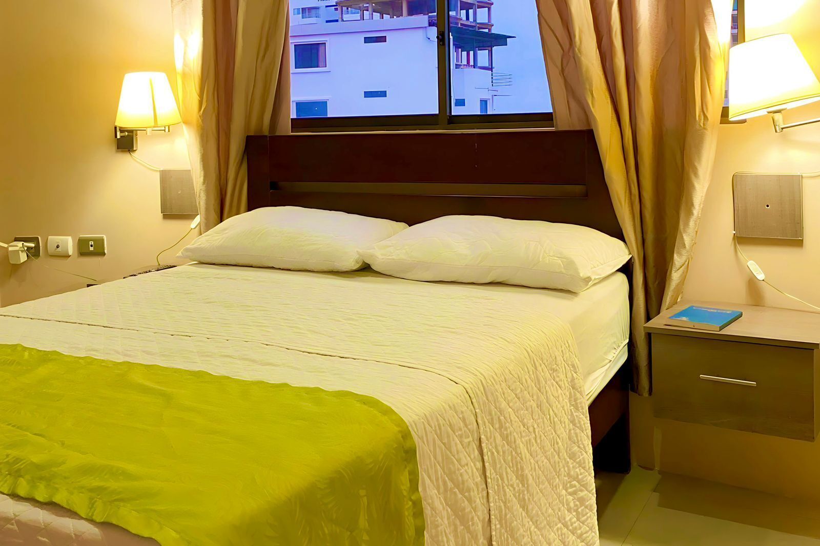 Habitación de hotel con cama, mesita de noche con un libro, lámparas de pared y cortinas en la ventana.