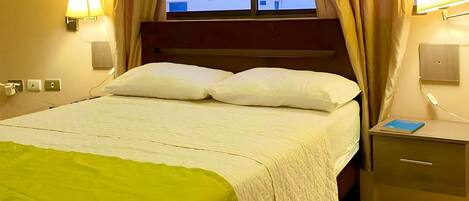Habitación de hotel con cama, mesita de noche con un libro, lámparas de pared y cortinas en la ventana.