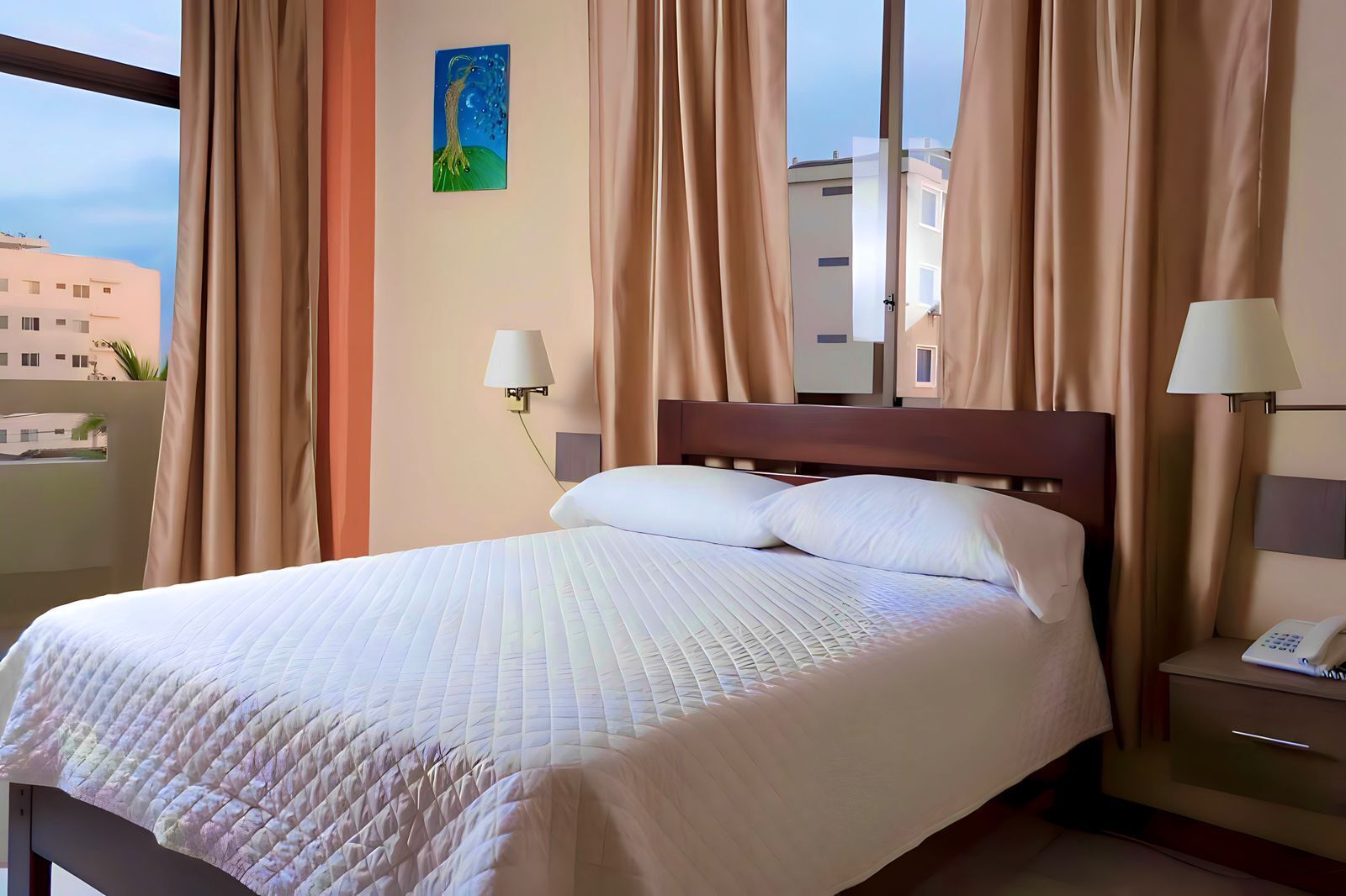 A hotel room with a large bed, beige curtains, a nightstand with a lamp, and a painting on the wall.