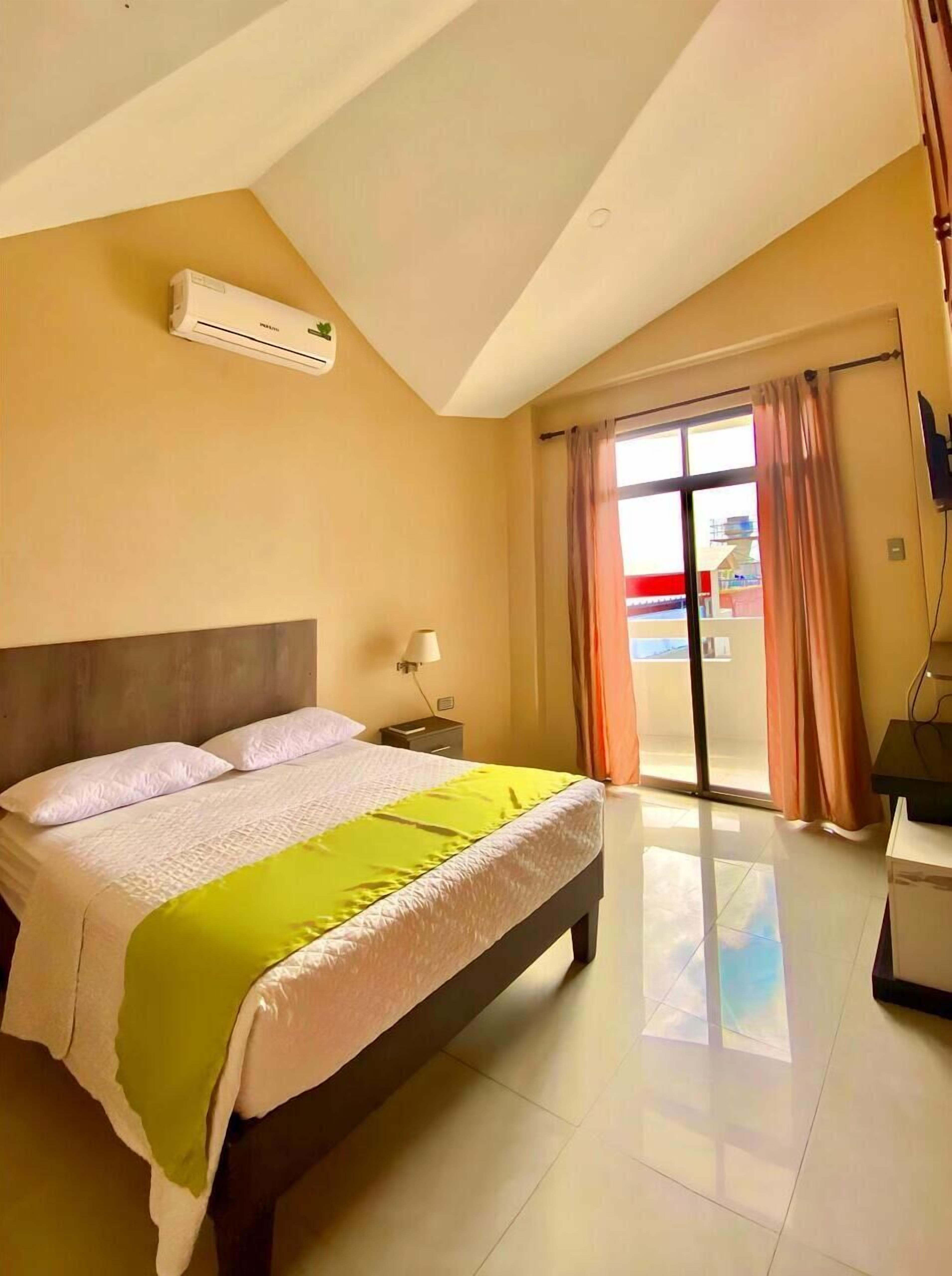 Una habitación de hotel con una cama grande, una mesita de noche, una lámpara y vistas al exterior a través de una puerta corrediza de vidrio.