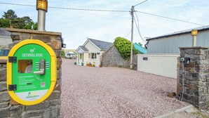 A defibrillator mounted on a stone wall, a gravel driveway, a small house with a blue roof, and a stone wall with a metal gate. - 1 bedroom accommodation in Ammanford (Ammanford)