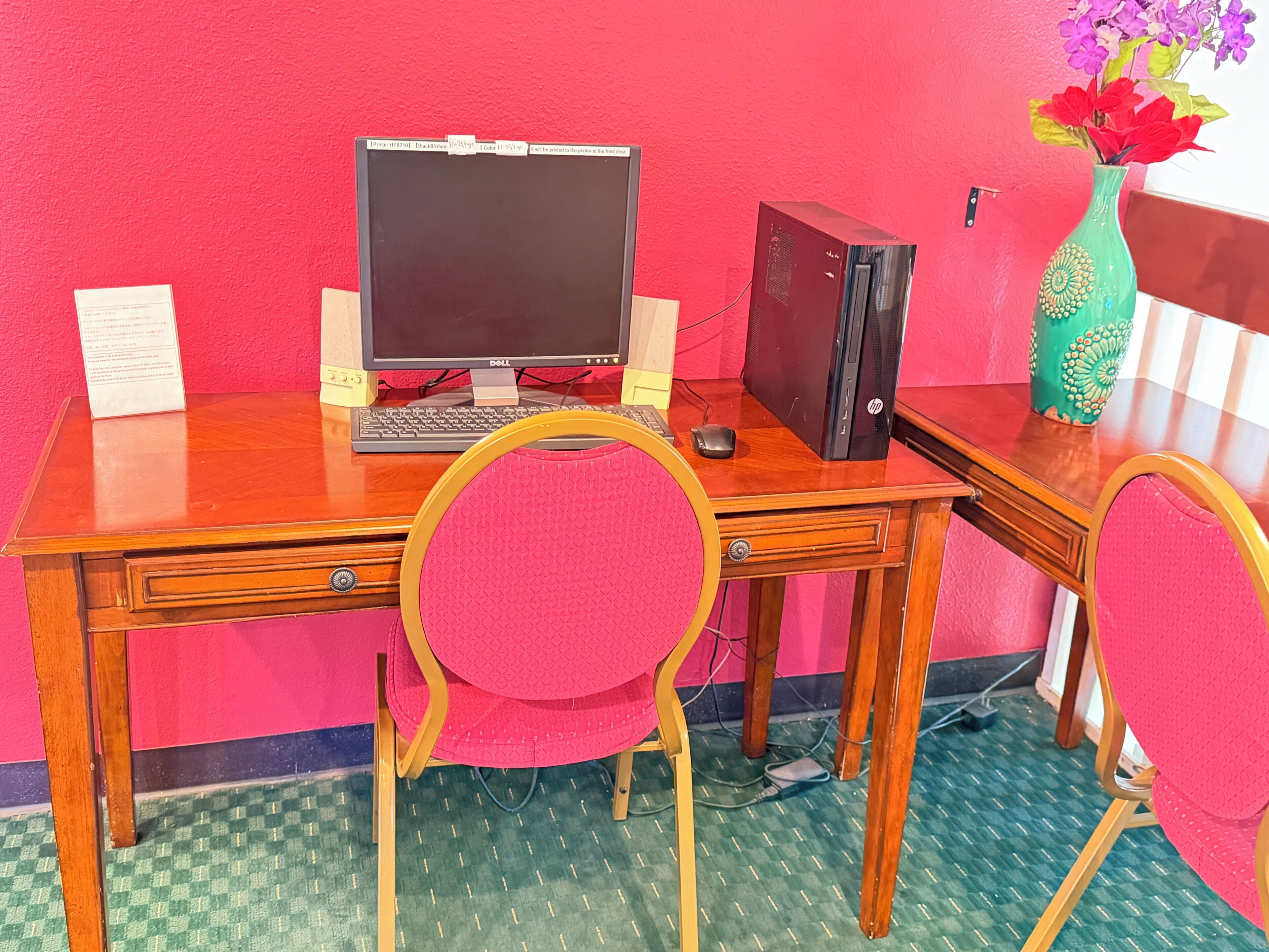 Un escritorio de madera con una computadora, una silla rosa y un florero con flores.