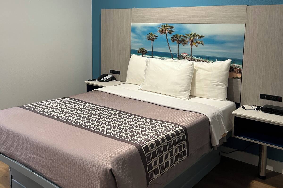 A hotel room with a bed, bedside tables, a nightstand, a painting of a beach scene, and a ceiling light.