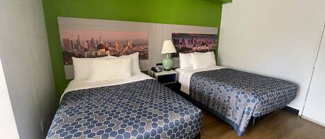 A hotel room with two beds, each with a patterned bedspread, a nightstand with a lamp, and a cityscape painting on the wall.