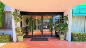 Entrada de hotel con suelo de baldosas, puertas de vidrio, plantas en macetas y un toldo verde.
