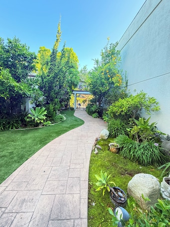 A paved pathway leading to a covered outdoor seating area surrounded by lush greenery and various plants.