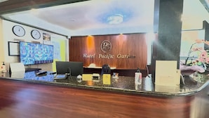 Un mostrador de recepción de hotel con un fondo de madera, un reloj, un televisor que muestra varias habitaciones y un letrero que dice 'Hotel Pacific Garden'.