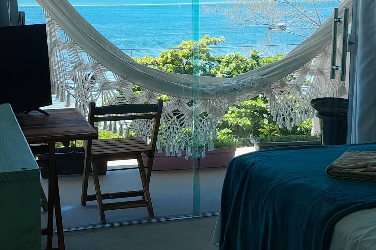 Quarto com cama, escrivaninha, cadeira e vista para o oceano através de uma porta de vidro.