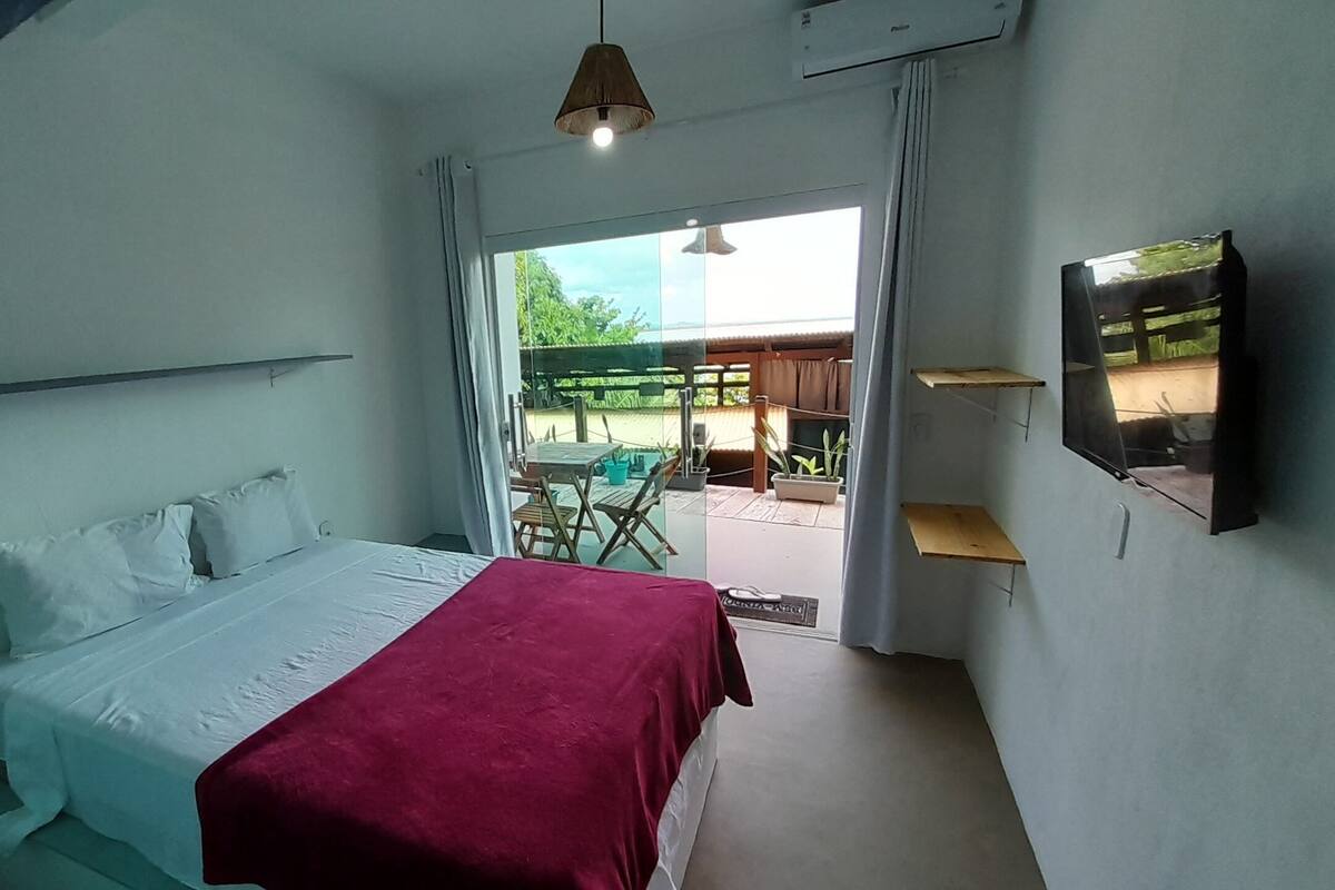 Um quarto com uma cama, um cobertor vermelho, um ventilador de teto, uma TV e vista para um pátio externo com uma mesa e cadeiras.