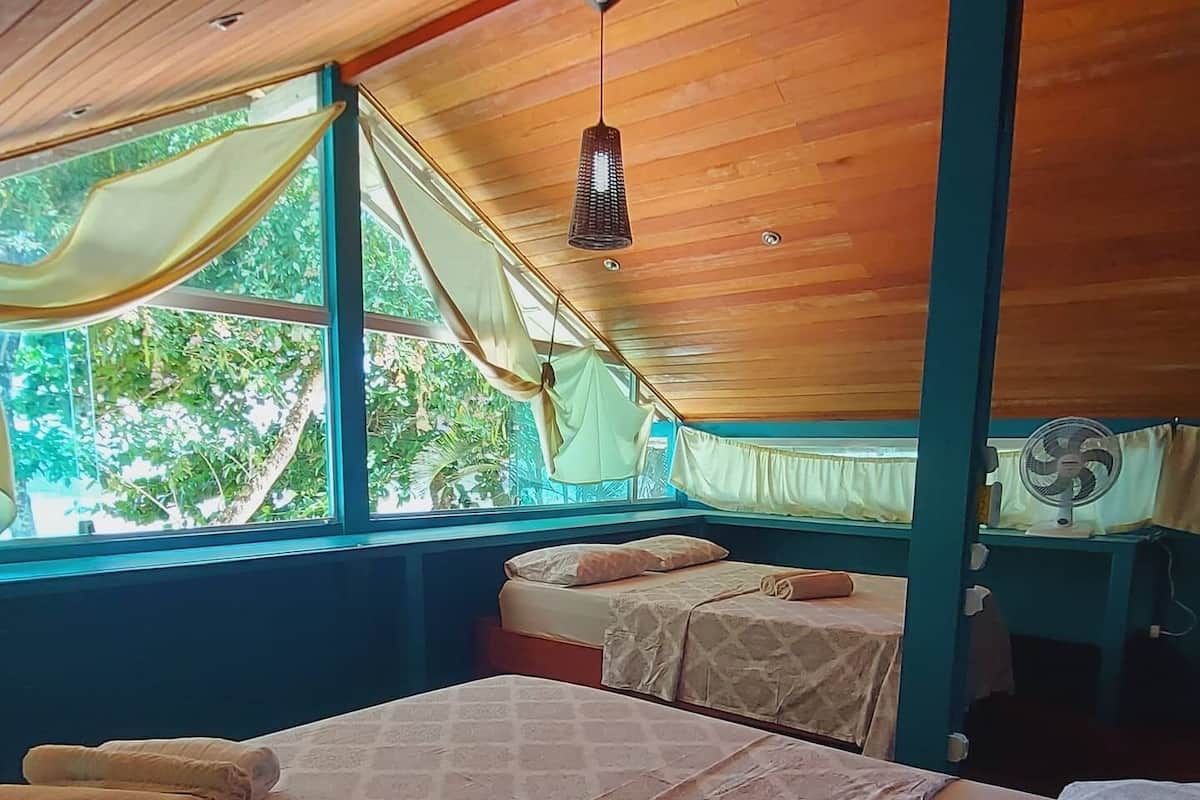 Quarto com duas camas, janela com cortinas, ventoinha no teto e vista para o verde lá fora.