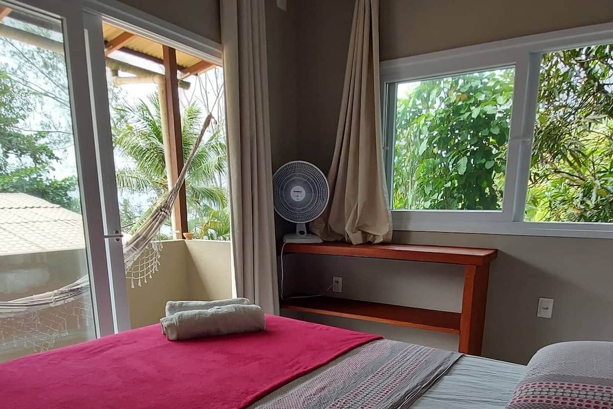Quarto com cama, ventilador e janela com vista para vegetação.