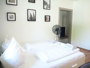 A neatly made bed with white linens, a standing fan, and framed pictures on the wall.