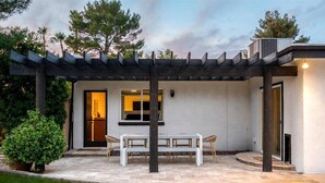 En moderne udendørs terrasse med et pergolaoverdækning, et bord med stole og en velholdt græsplæne.