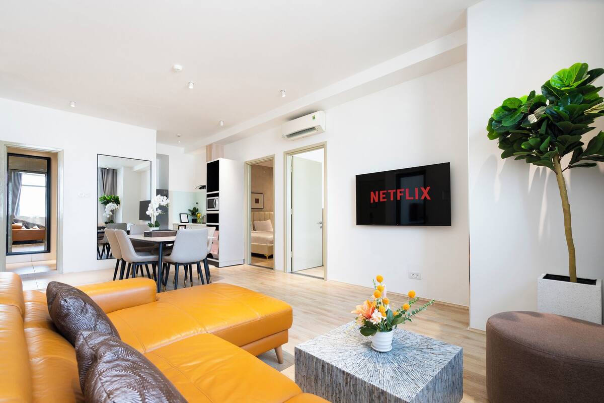 Ein modernes Wohnzimmer mit einer großen Couch, einem Couchtisch mit Blumen, einem an der Wand montierten Fernseher und einem Essbereich im Hintergrund.