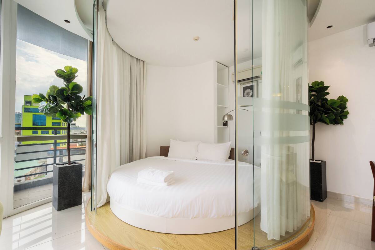 Ein modernes Schlafzimmer mit einem runden Bett, einer Glastrennwand und einem Balkon mit Blick auf Gebäude.