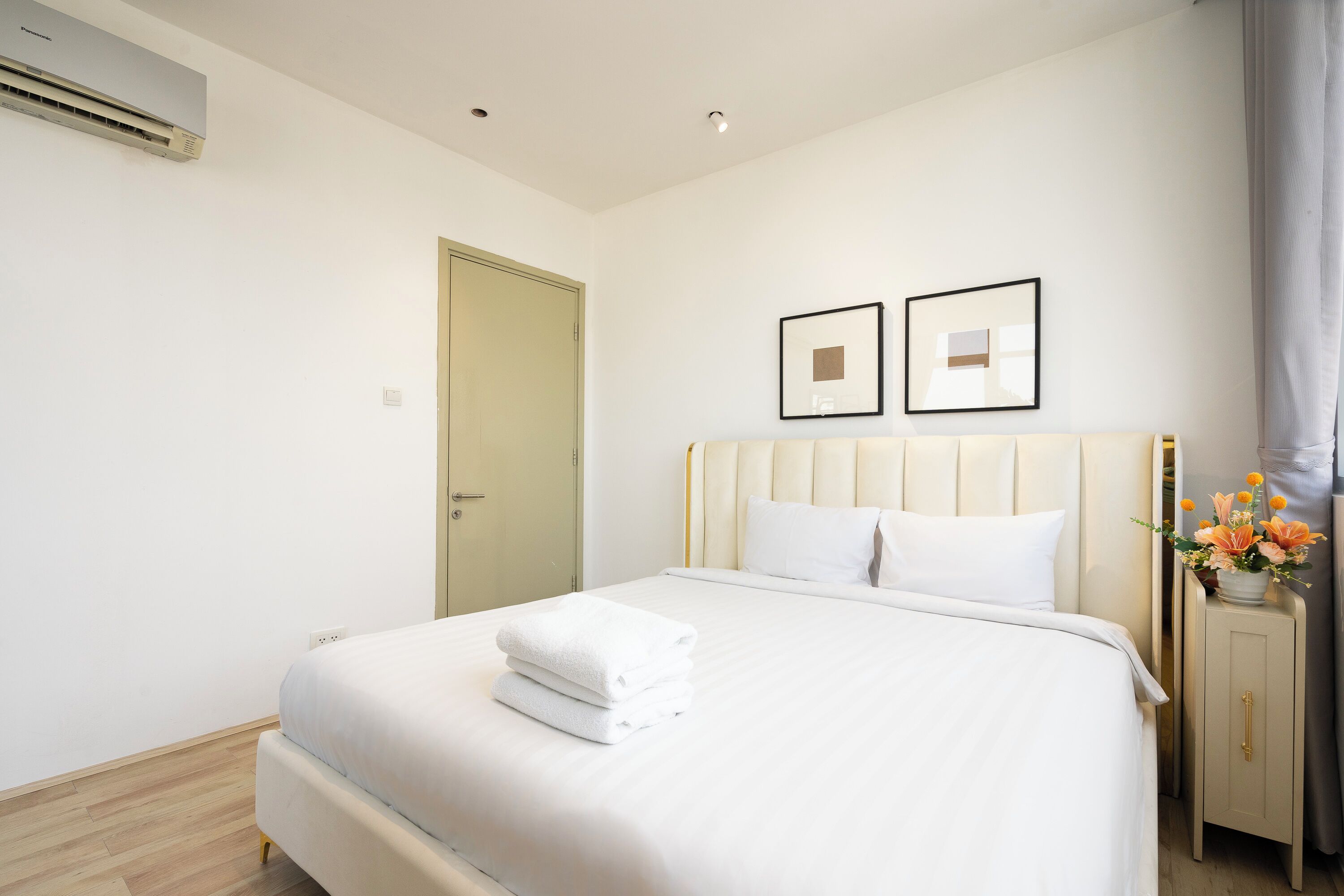 A neatly made bed with white linens and a folded towel, a bedside table with a vase of flowers, and a door to the left.