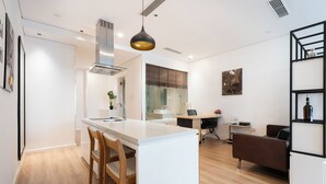 A modern kitchen with a central island, wooden chairs, a dining table, a sofa, and a black shelving unit. - THE ONE Saigon Luxury Citypoint By An Gia Hospitality (Ho Chi Minh City)