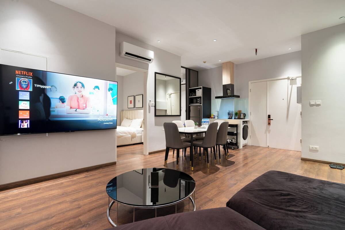 Un salon moderne avec un téléviseur à écran plat, une table basse ronde et un canapé. Il y a également un coin repas avec une table et des chaises. La pièce est dotée d’un parquet et de murs blancs.