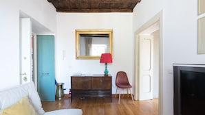 Une pièce avec un plancher en bois, un canapé blanc avec des coussins, une lampe rouge, un meuble en bois, une chaise rouge, un grand miroir et un téléviseur.