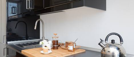 A modern kitchen with a stainless steel kettle, a coffee maker, and a wooden cutting board with food items.