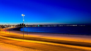 A nighttime beach scene with a road leading to the water, streetlights, and distant city lights. - Luxurious modern apartment in Reykjavik with ocean view! (Reykjavík)