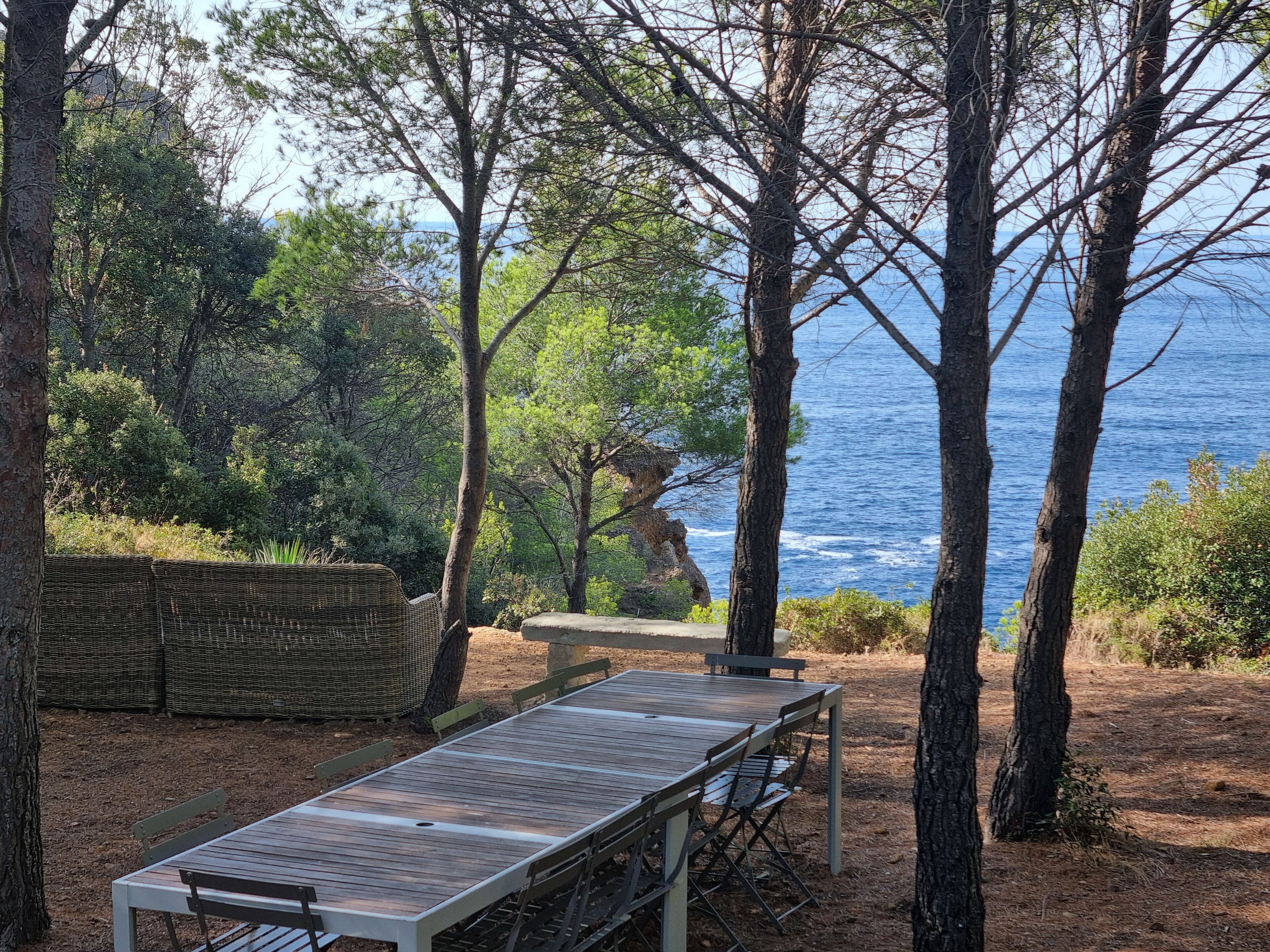 An outdoor seating area with a wooden table and chairs, surrounded by trees and overlooking a body of water.