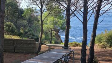An outdoor seating area with a wooden table and chairs, surrounded by trees and overlooking a body of water.