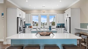 A modern kitchen with white cabinets, a large island with stools, stainless steel appliances, and a view of the neighborhood through large windows. - New luxury ocean front w/Heated private pool, Elevator, 8 Bed/8 Bath (Holden Beach)