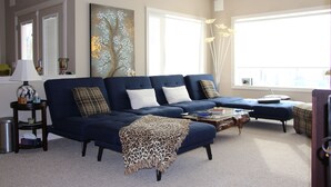 A living room with a dark blue sofa, a coffee table, and a leopard print throw. There is a painting on the wall, a lamp on a side table, and a window with blinds. - Charming Guest House with Beautiful Mountain View & Spa....Pet Friendly (De Winton)