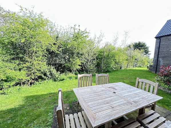 Une table et des chaises en bois sont disposées dans un jardin, avec un bâtiment en briques en arrière-plan.